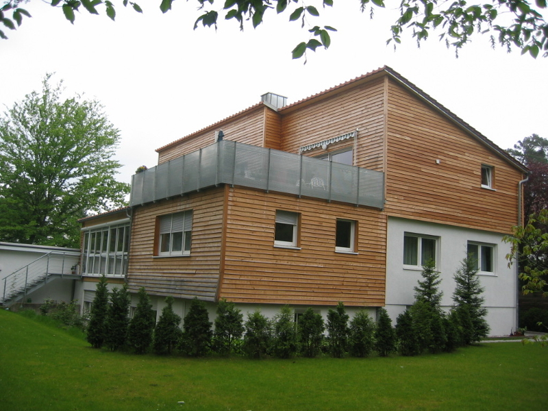 Ein modernes zweistöckiges Haus mit Holzfassade, weißem Anbau und Terrasse, umgeben von Grünflächen und Bäumen.